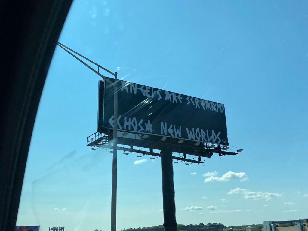 Picture taken out of the window of a moving car on a clear day. Outside the window is a billboard with large white letters on a black background that reads: "ANGELS ARE SCREAMS ECHOS A NEW WORLDS"