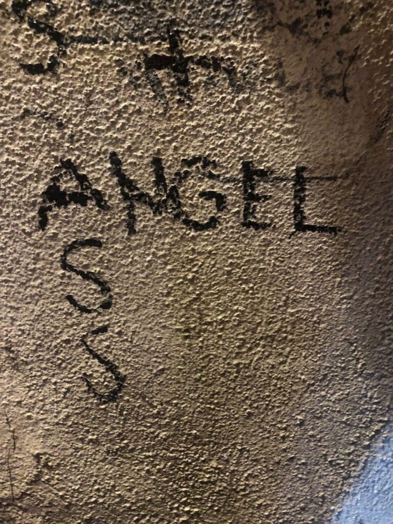 Close up of a stucco wall, where someone has written an acrostic "Angel, Ass" where the "A" is shared between words.