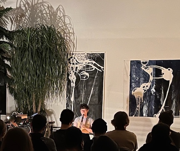 Clarinetist Madison Greenstone performs 'exstatic resonances' for a seated roomful of audience members in front of large ink-prints and a large potted indoor tree. The audience listens attentively.