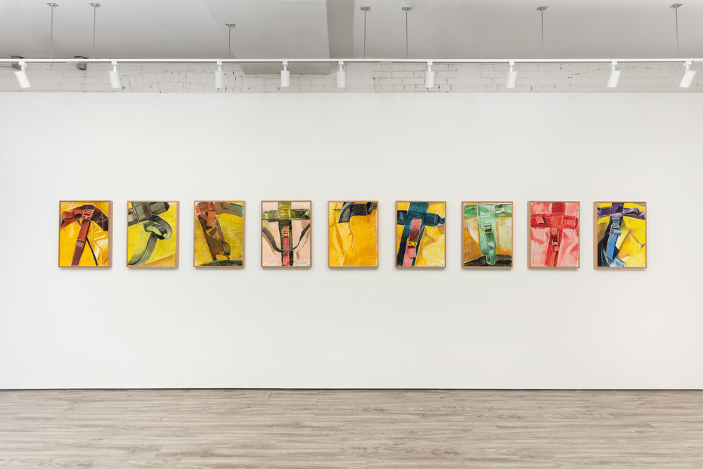An exhibition installation view of 9 framed bold colored pastel drawings of a close-up view of a duffel bag. Each drawing shows criss crossing straps on a loosely drawn background.